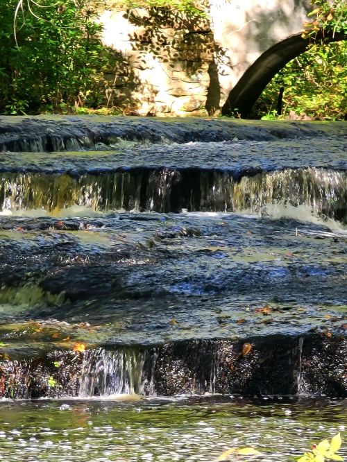 A serene waterfall cascades over rocky steps, surrounded by lush greenery and a stone bridge in the background.