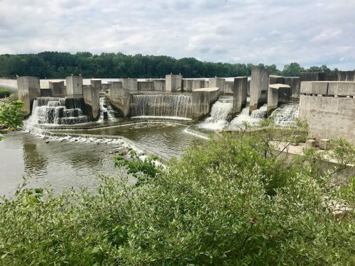 A concrete dam with cascading water, surrounded by greenery and a cloudy sky in the background.