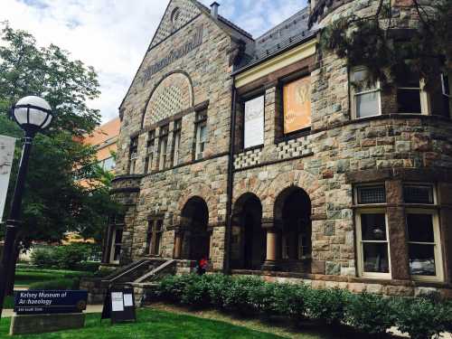 Stone building of the Kelsey Museum of Archaeology, featuring large windows and a sign in front, surrounded by greenery.