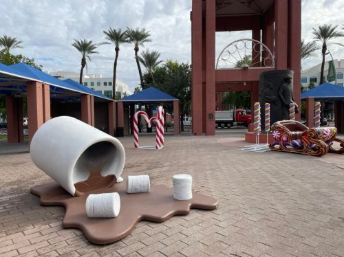 A large, tipped-over cup spilling chocolate, with marshmallows and candy canes, set in a festive outdoor area.