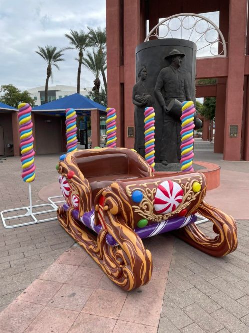 A colorful, oversized candy-themed sleigh with lollipop decorations, set in a plaza with palm trees in the background.