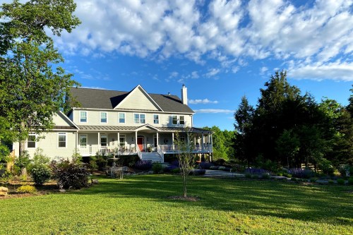 A large, two-story house with a porch, surrounded by lush green grass and trees under a blue sky with scattered clouds.