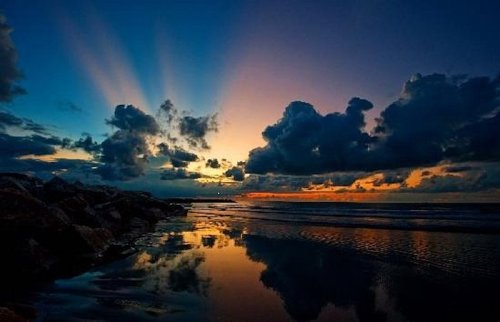 A serene beach at sunset, with colorful clouds reflecting on the water and rays of light breaking through the sky.