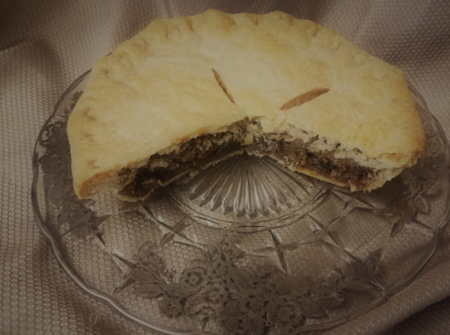 A round pie with a golden crust, partially sliced, revealing a dark filling, displayed on an ornate glass plate.