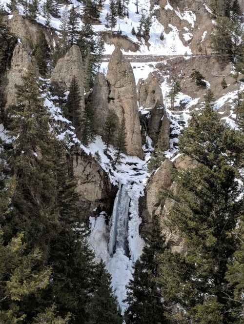 A frozen waterfall cascades between rocky cliffs, surrounded by snow-covered trees and rugged terrain.