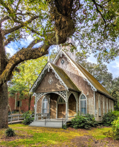 A charming wooden church with intricate architecture, surrounded by lush greenery and large trees under a blue sky.