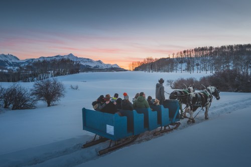 A group of people in a blue sleigh pulled by horses, enjoying a winter sunset in a snowy landscape.