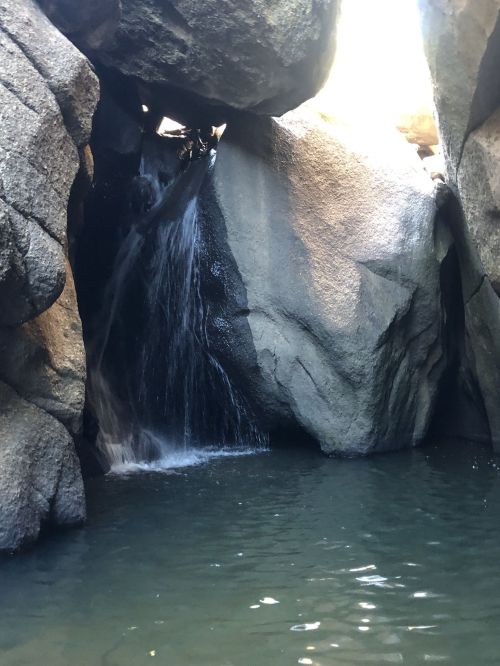 A small waterfall cascades between large rocks into a tranquil pool of water. Sunlight filters through the opening above.