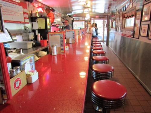 A retro diner interior featuring a long red counter with stools and a few patrons, framed by vintage decor.