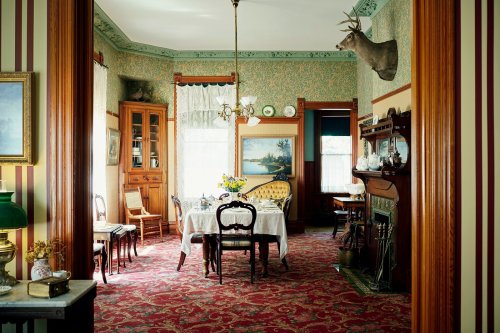 A vintage dining room with floral wallpaper, a table set for meals, and a mounted deer head on the wall.