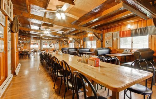 Cozy wooden interior of a restaurant with a long table, black chairs, and rustic decor. Bright windows let in natural light.