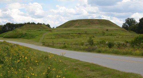 Two large grassy mounds rise in the distance, flanked by a winding road and wildflowers under a cloudy sky.