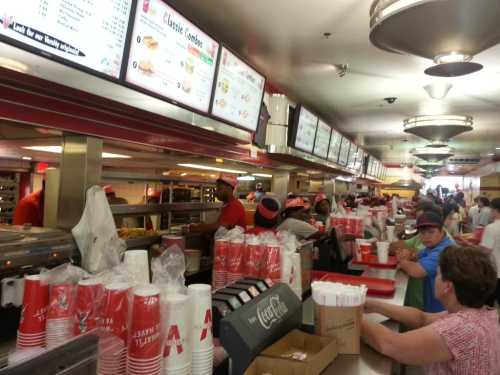 A busy fast-food restaurant with a long counter, staff serving food, and customers waiting in line. Red and white decor.