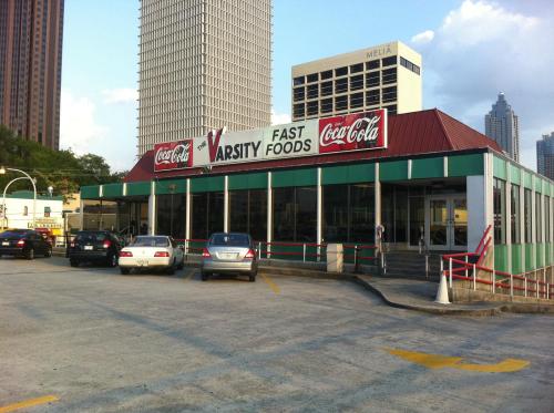 Exterior of Varsity Fast Foods restaurant with Coca-Cola signage, surrounded by parked cars and city buildings in the background.