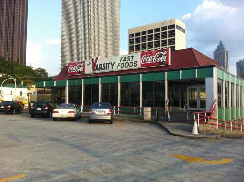 Exterior of Varsity Fast Foods restaurant with Coca-Cola signage, surrounded by parked cars and city buildings in the background.