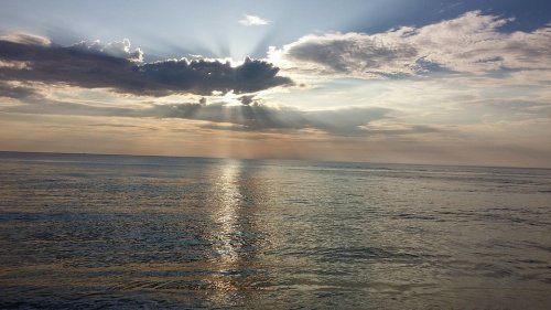 Sunlight breaks through clouds over a calm ocean, creating a serene reflection on the water's surface.