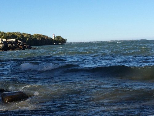 Waves gently roll in on a lake, with a distant lighthouse and trees visible on the shore under a clear blue sky.