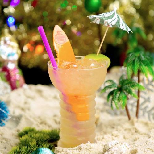 A colorful tropical cocktail in a fun cup, garnished with a mini umbrella and a surfboard, set on sandy decor.