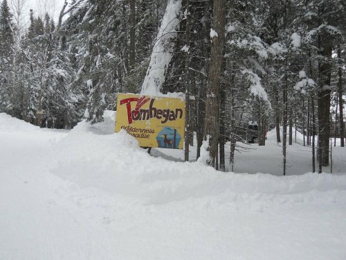 A colorful sign for Tomhegan, partially buried in snow, surrounded by tall trees in a winter landscape.