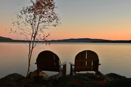 Two wooden chairs facing a calm lake at sunset, with a lone tree nearby and soft pastel colors in the sky.