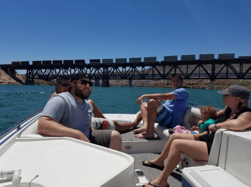 A group of people relax on a boat, with a train bridge in the background and clear blue water around them.