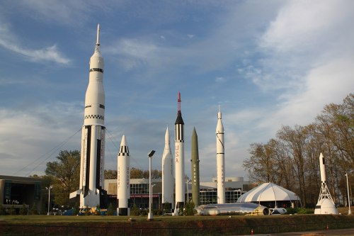 A collection of various rockets displayed outdoors, with trees and a building in the background under a cloudy sky.
