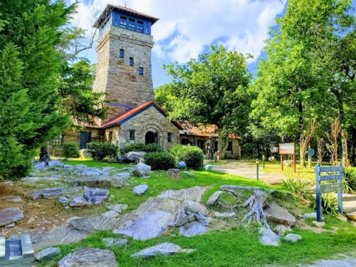 A stone tower and building surrounded by trees and rocks under a blue sky, with a path leading to the entrance.