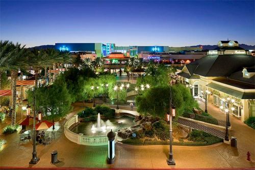 A vibrant outdoor shopping area at dusk, featuring palm trees, a fountain, and colorful lights illuminating the scene.