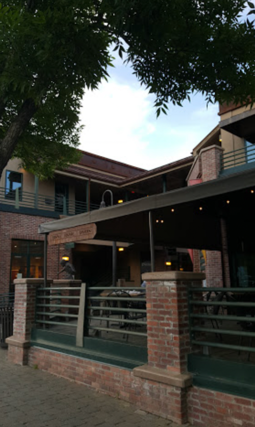 A brick building with a covered patio, surrounded by trees and a clear sky. Outdoor seating is visible.