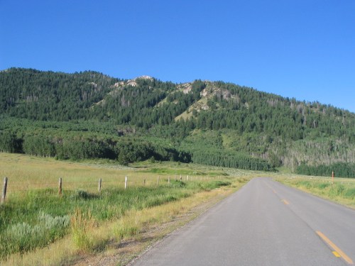 A scenic road leads through a green valley, flanked by trees and mountains under a clear blue sky.