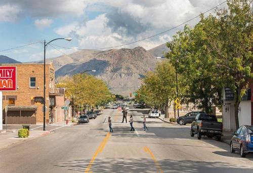 A small town street with pedestrians crossing, surrounded by trees and mountains under a partly cloudy sky.