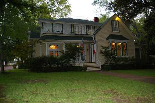 A charming two-story house with a porch, surrounded by greenery, featuring decorative windows and flags.