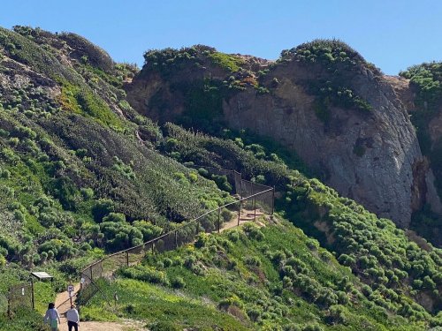Two people walk along a path beside a green hillside with a fence and rocky cliffs under a clear blue sky.