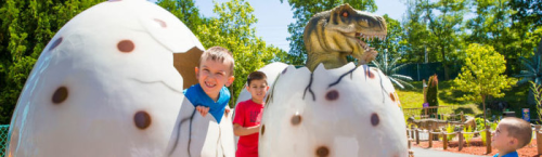 Two children peek out from large dinosaur eggs, with a dinosaur figure visible in the background on a sunny day.