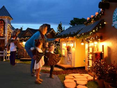 A woman and children admire a decorated miniature house with festive lights at dusk.