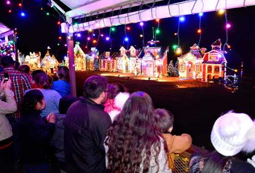 A crowd watches a colorful display of illuminated holiday houses decorated with lights at night.