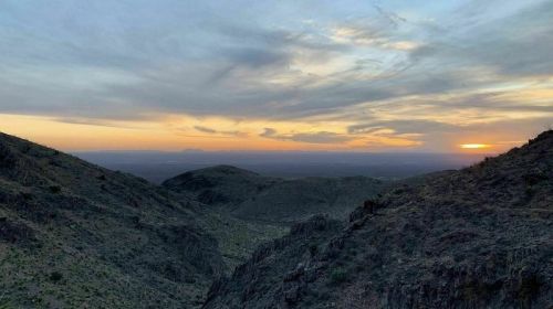 A scenic view of rolling hills at sunset, with a colorful sky and sparse vegetation in the foreground.