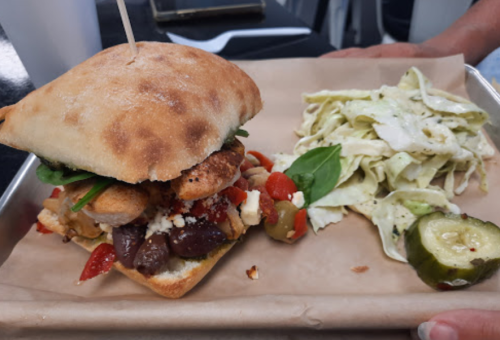 A sandwich with grilled chicken, vegetables, and feta, served with coleslaw and a pickle on the side.