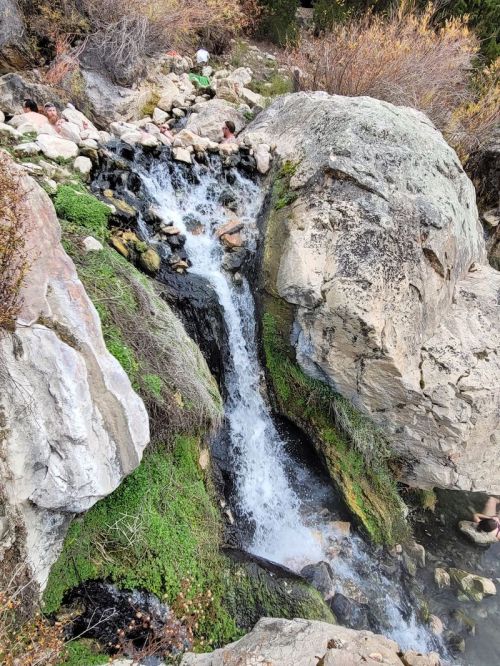 A small waterfall cascades over rocks, surrounded by greenery and boulders in a natural setting.