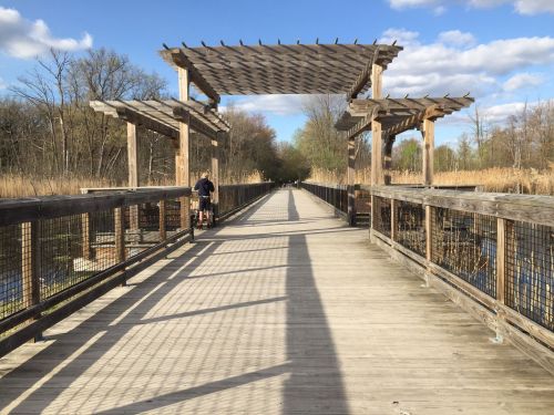 A wooden boardwalk leads through a natural area, framed by a pergola and surrounded by tall grasses under a blue sky.