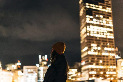 A person in a beanie stands against a city skyline at night, with illuminated buildings and a cloudy sky.