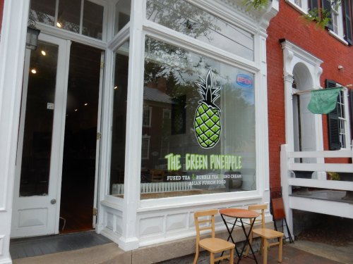 A storefront with a large window displaying "The Green Pineapple" sign, featuring a pineapple graphic and outdoor seating.