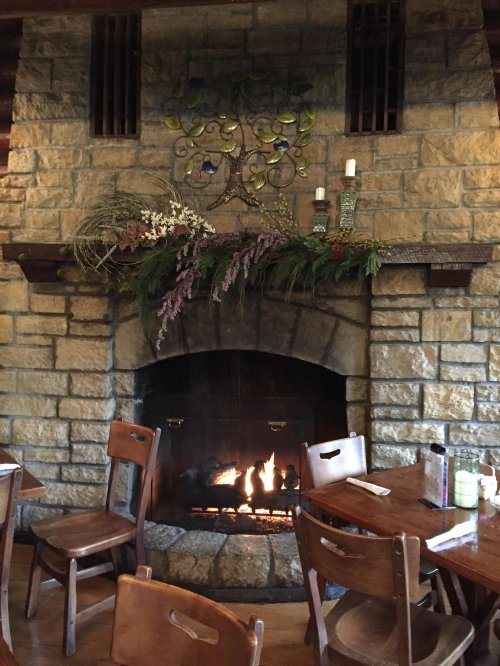 Cozy restaurant interior featuring a stone fireplace adorned with greenery and flowers, surrounded by wooden tables and chairs.