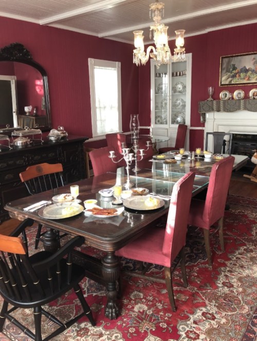 A dining room with a dark wooden table set for breakfast, surrounded by red chairs and a chandelier above.