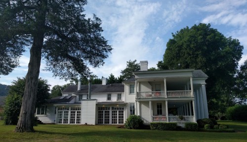 A large white house with multiple stories, surrounded by green trees and a grassy lawn under a blue sky.