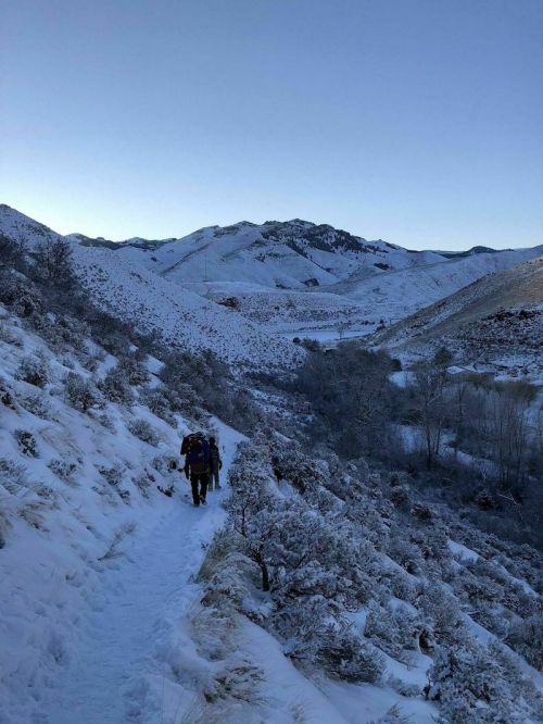 Two hikers walk along a snowy trail in a mountainous landscape under a clear blue sky.