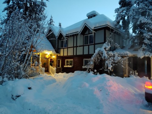 A cozy, snow-covered house with warm lights glowing at dusk, surrounded by tall trees and a thick blanket of snow.