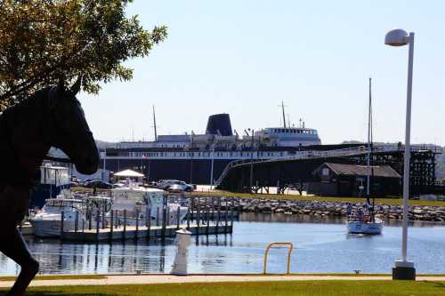 A marina with boats and a large ship in the background, framed by trees and a horse statue in the foreground.