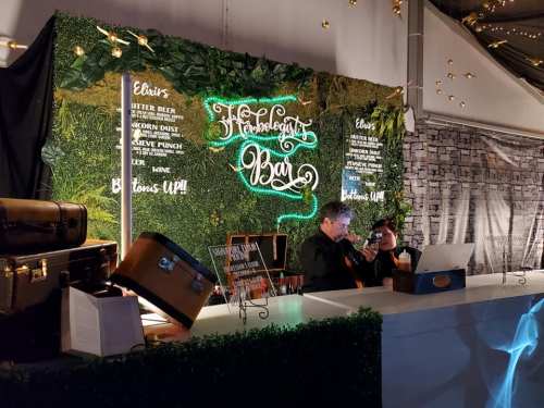 A vibrant bar setup with greenery, neon sign reading "Hobbledehoy Bar," and two people enjoying drinks.