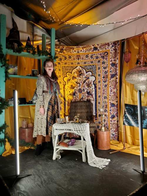 A woman stands in a decorated space with colorful fabrics, a table, and various decorative items.